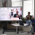 Uczestniczka prezentuje podczas PHD RISK Summerschool, obok ekran z informacją o warsztatach, w nowoczesnym biurze z dużymi oknami i naturalnym oświetleniem.