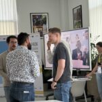 Uczestnicy PHD RISK Summerschool rozmawiają podczas wydarzenia, prezentacja na ekranie i materiały konferencyjne w tle, w nowoczesnym biurze.