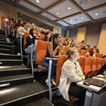 Grupa studentów i naukowców uczestniczących w PHD RISK Summerschool podczas wykładu w nowoczesnej sali konferencyjnej.