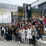 Grupa uczestników PHD RISK Summer School przed budynkiem Uniwersytetu Gdańskiego, z logo uczelni i nowoczesną architekturą w tle, podczas wydarzenia naukowego.