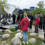 Grupa uczestników PHD RISK Summer School na zewnątrz, obserwująca instalację na moście nad wodą, w otoczeniu zieleni i nowoczesnych budynków w tle.